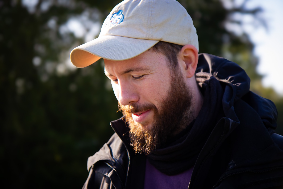Ein bärtiger Typ, der eine schwarze Regenjacke, ein lila Shirt und eine beige Cappy trägt. Sein Blick ist nach unten gerichtet. Im weichgezeichneten Hintergrund ist die Natur zu sehen.